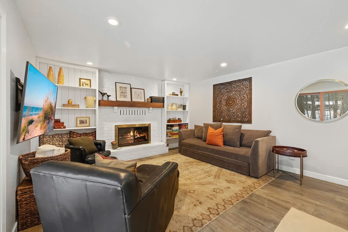 A cozy living room features a white brick fireplace flanked by built-in bookshelves, a large brown sofa, and two dark leather armchairs, all centered around a patterned rug.