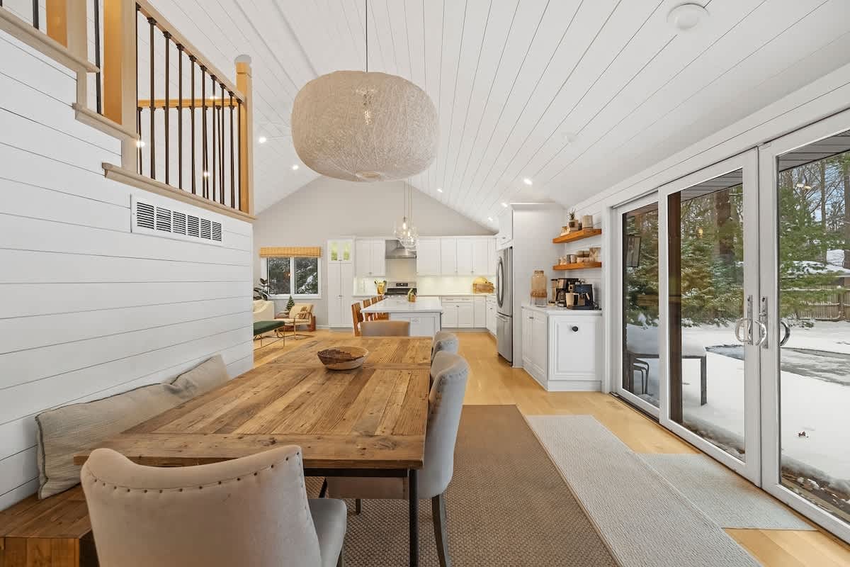 Rustic dining area with a large wooden table and upholstered chairs, illuminated by a large spherical pendant light, adjacent to a white shiplap wall and sliding glass doors leading to a snowy exterior.