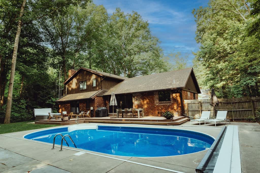 A two-story house with brick and wood siding overlooks a large kidney-shaped swimming pool, surrounded by a concrete deck with lounge chairs and a wooden deck with outdoor dining, all nestled among tall green trees.