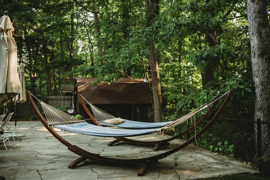 Two comfortable hammocks with wooden stands are set on a flagstone patio, nestled among a dense forest of green trees, creating a serene outdoor relaxation spot.