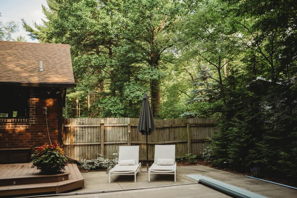 Serene backyard area featuring a wooden fence, lush greenery, and two white lounge chairs with an umbrella on a paved section next to a wooden deck.