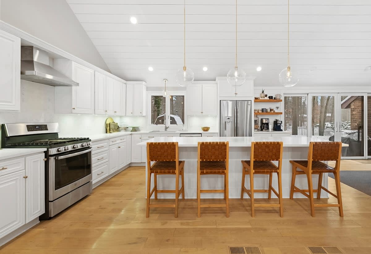 Modern white kitchen with stainless steel appliances, a large island with four woven bar stools, and three clear glass pendant lights, set against a vaulted shiplap ceiling and hardwood floors.