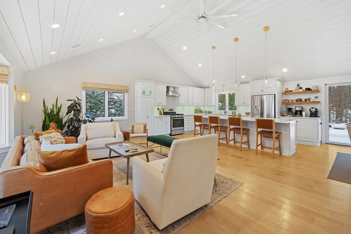 Open concept living space featuring a vaulted shiplap ceiling, a cozy living area with an orange sofa and cream armchairs, flowing into a modern white kitchen with an island and bar stools.