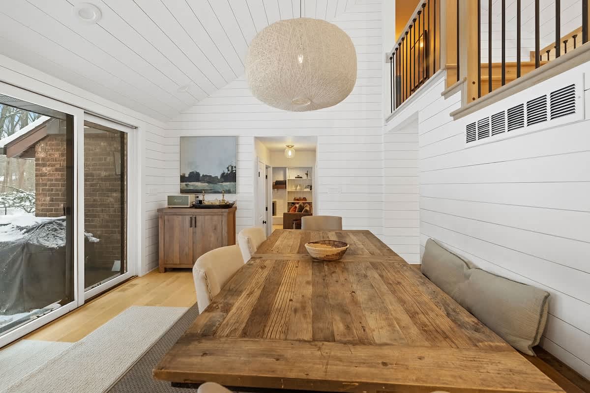 Dining area featuring a rustic wooden table, a large spherical pendant light, and a shiplap accent wall with abstract art, offering a glimpse into a hallway and outdoor views through sliding glass doors.