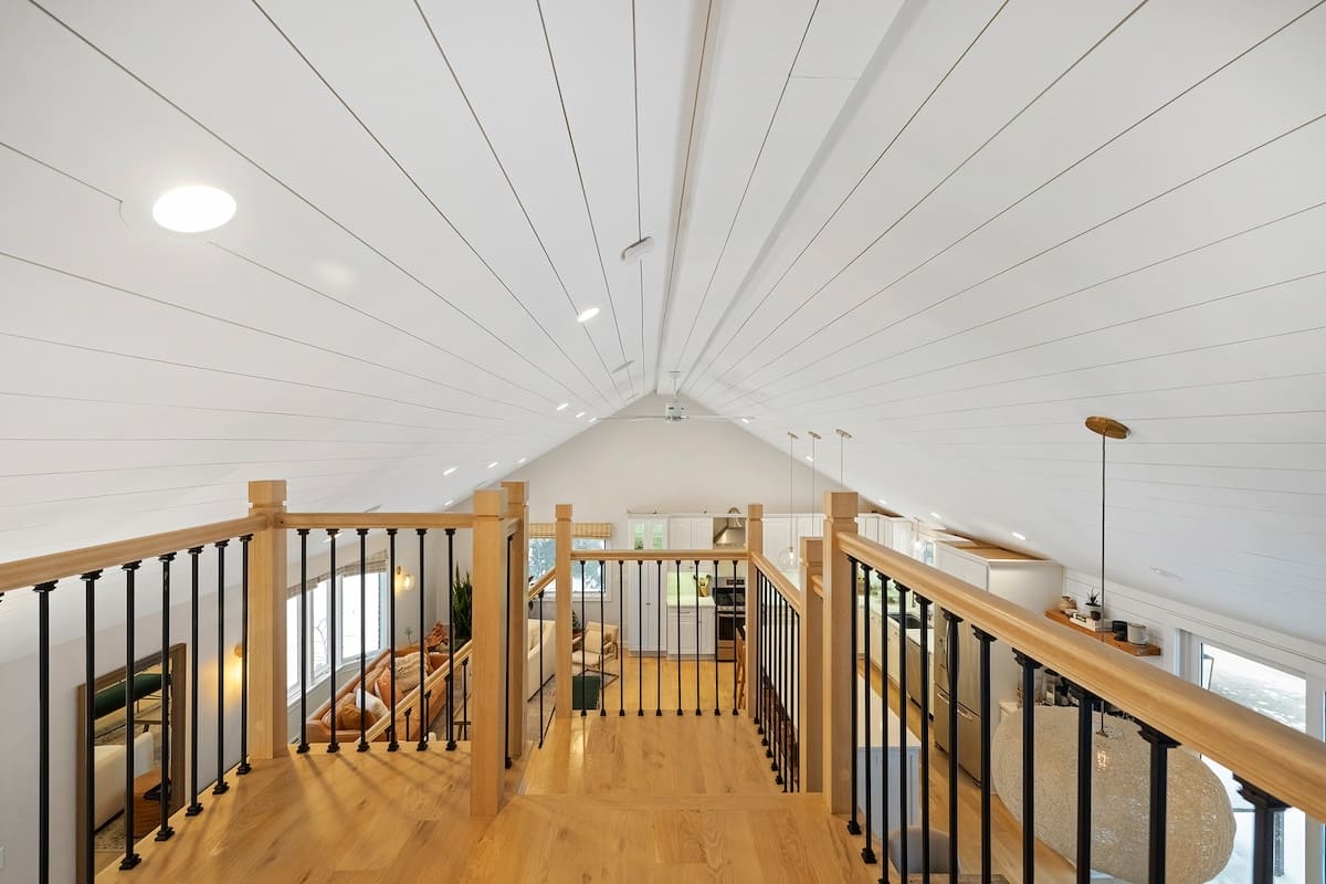 View from a wooden loft with black metal railings, looking down into a spacious open-concept living area with vaulted white shiplap ceilings and light hardwood floors.