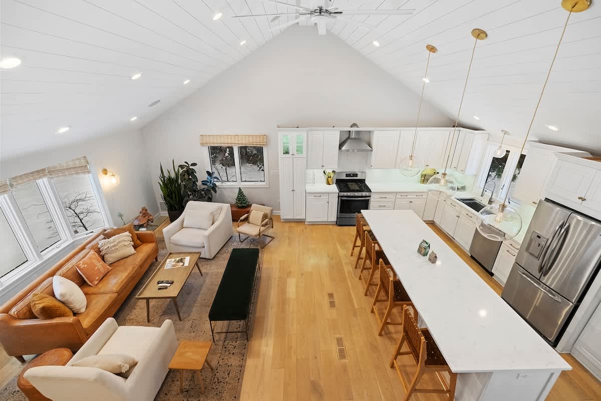 Expansive open-concept living room and kitchen with vaulted white shiplap ceilings, light hardwood floors, and large windows. The living area features a leather sofa and armchairs, while the kitchen has white cabinetry and a long island with seating.