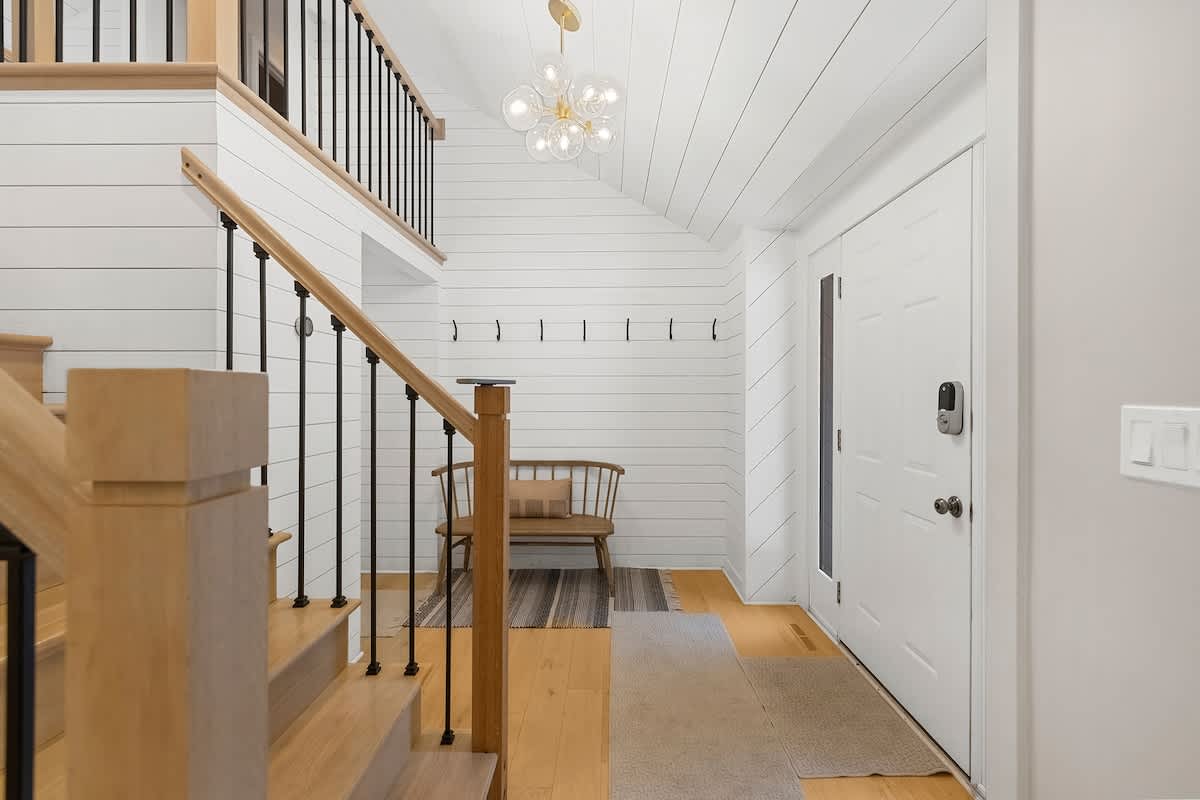 A bright entryway features white shiplap walls and ceiling, a modern chandelier, a wooden bench, and a light wood staircase with black metal balusters, leading to the upper floor.