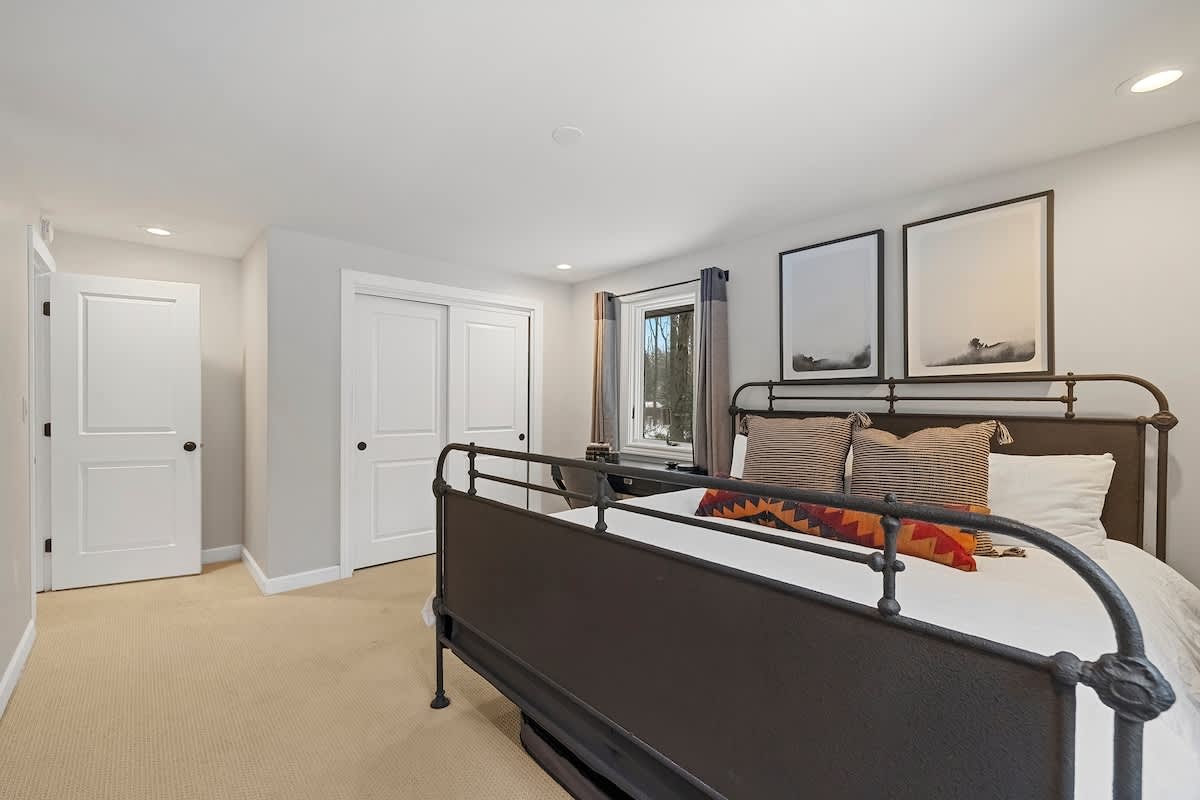 View of a bedroom with a dark metal bed frame and white bedding, showing a closet with sliding doors and a white door leading to another room. Two framed art pieces hang above the bed, and the room is carpeted.