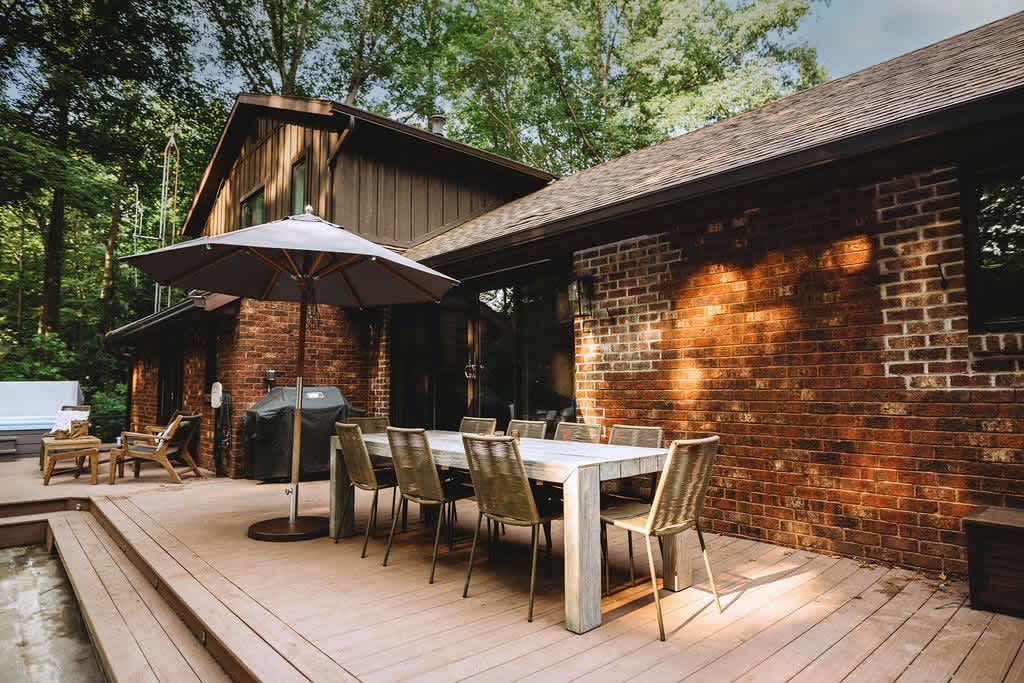 A spacious wooden deck features an outdoor dining table with chairs under a large umbrella, adjacent to a brick-sided house with a dark roof, surrounded by lush green trees. A BBQ grill is also visible.