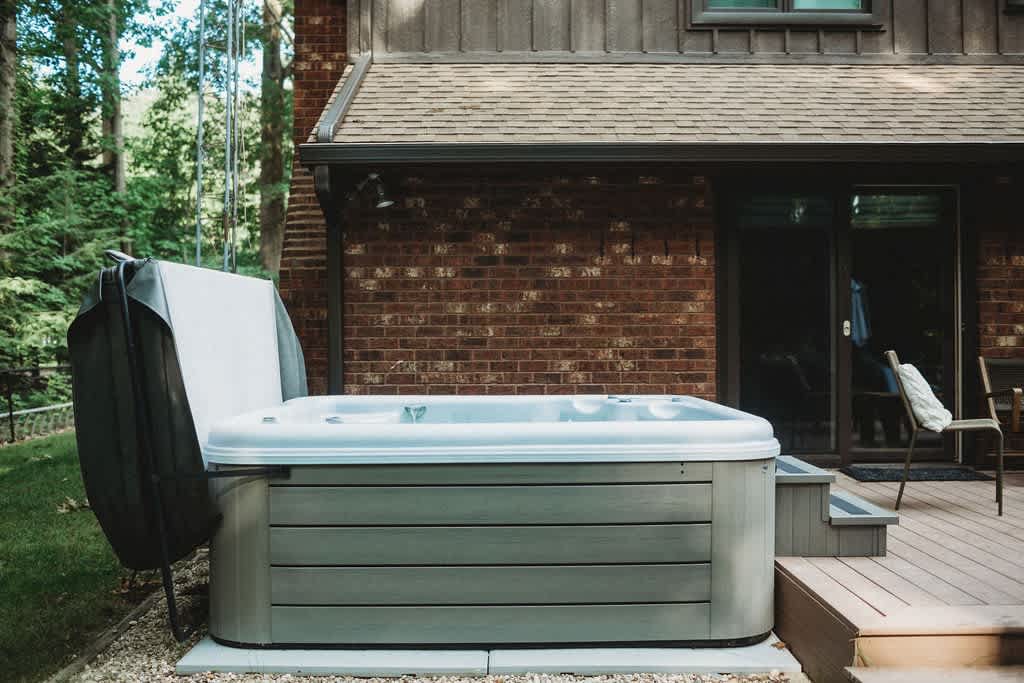 Modern grey hot tub with its cover partially open, situated on a gravel base next to a wooden deck and the brick exterior of the house.