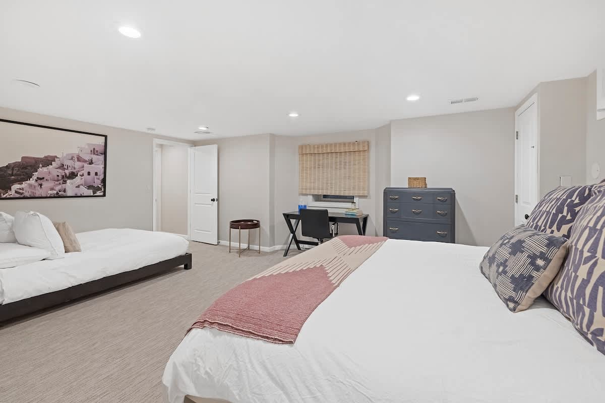 Expansive bedroom with two queen beds, white bedding, and decorative pillows, featuring a dark gray dresser and a desk area with a chair by a window. A large framed artwork hangs on the wall, and the room is carpeted.