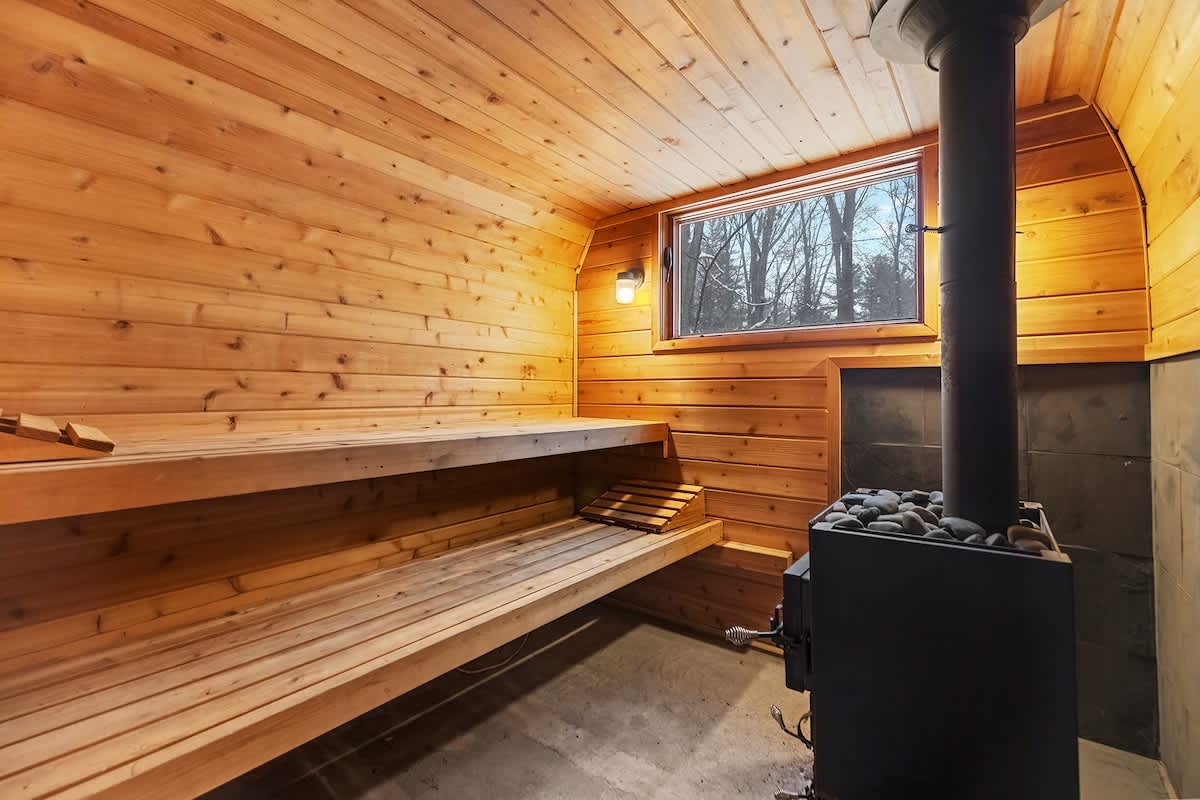A cozy wooden sauna features two tiers of benches, a window offering a view of trees, and a traditional wood-burning stove with rocks, creating a warm and inviting atmosphere.