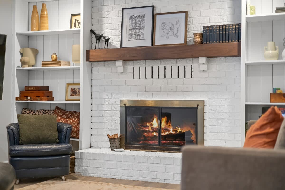 A detailed view of a white brick fireplace with a warm fire burning, topped by a rustic wooden mantel and flanked by white built-in bookshelves adorned with decorative items and framed artwork.