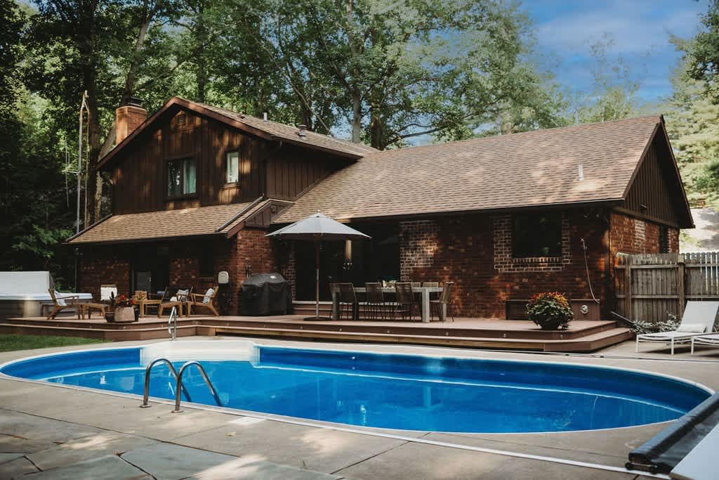 Traditional brick and wood-sided house with a large wooden deck, outdoor dining area, and a blue kidney-shaped swimming pool in the foreground. Mature trees surround the property.