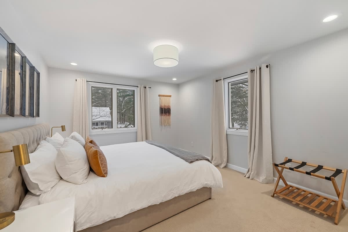 Cozy bedroom with a queen bed, white bedding, and a light gray throw, positioned between two large windows with light-colored curtains. A wooden luggage rack stands near one window, and a woven wall hanging adds texture to the light gray walls.