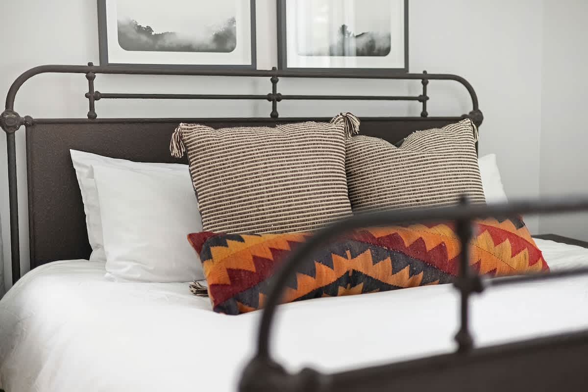 Close-up view of a bed with a dark metal frame, white sheets, and a variety of decorative pillows including striped and geometric patterns. Two framed black and white art prints are visible above the headboard.