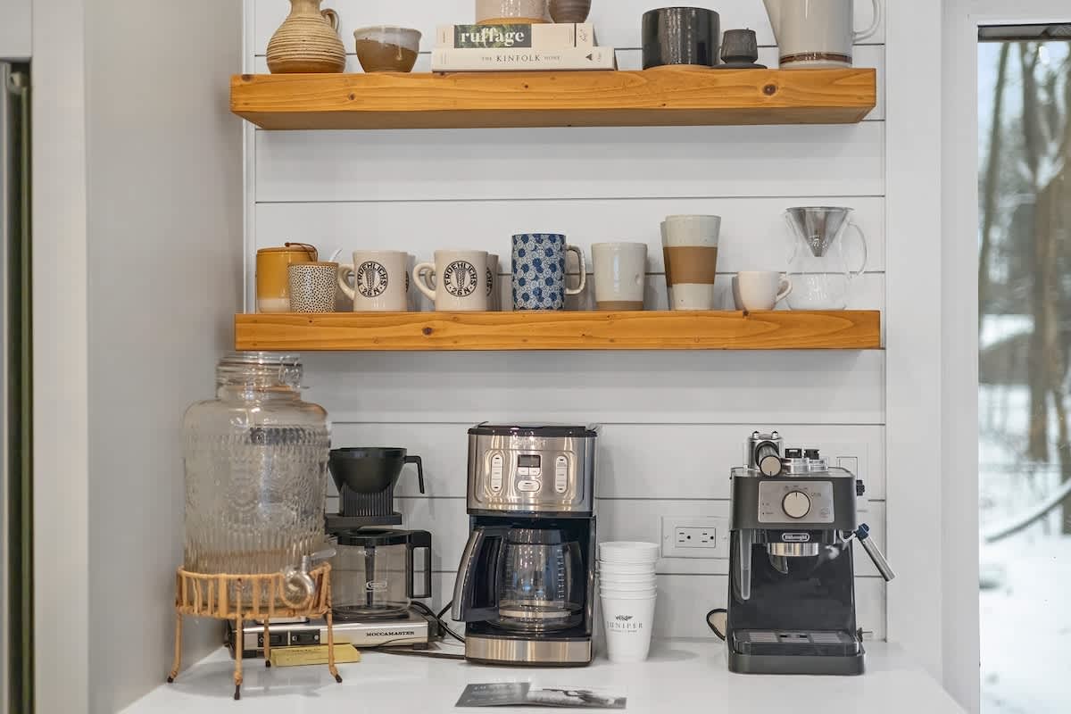 Dedicated coffee station in the kitchen, featuring wooden floating shelves stocked with mugs and decor, and a countertop with various coffee makers and a large beverage dispenser.