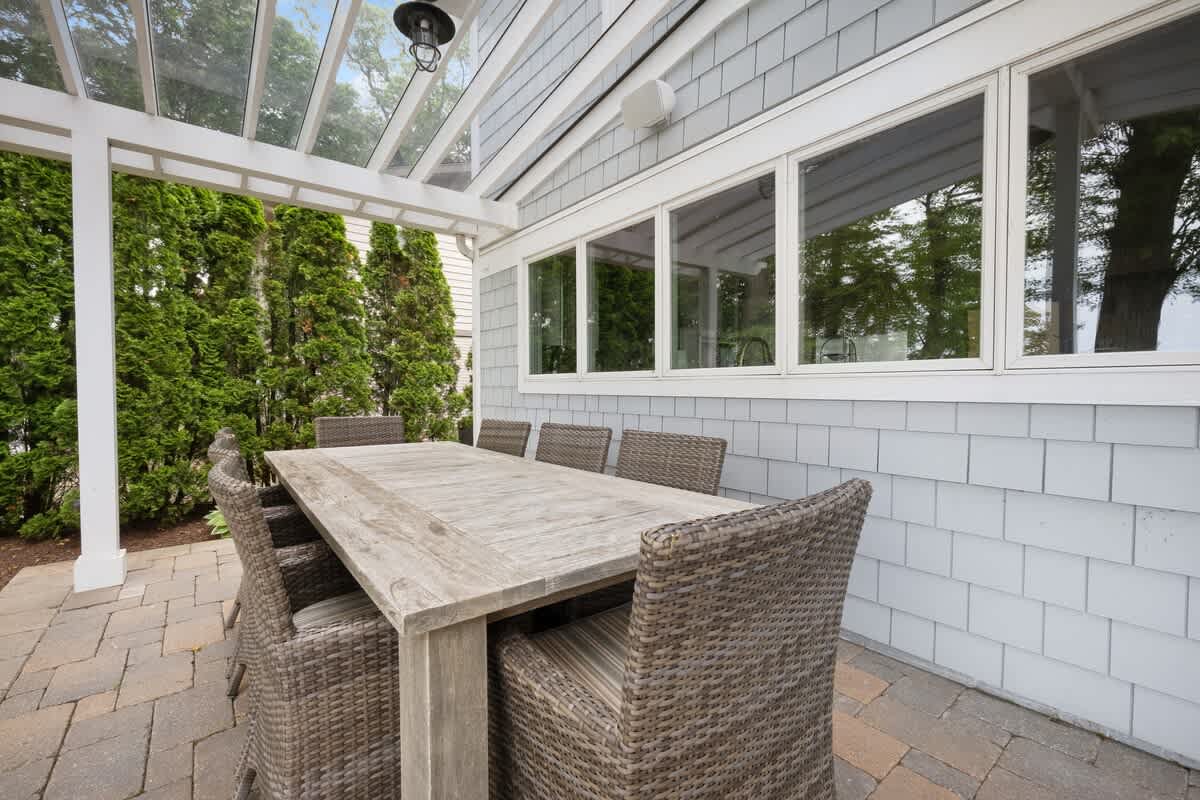 A rustic wooden dining table with wicker chairs sits on a paved patio, partially covered by a clear-roofed pergola, adjacent to the light gray shingle-sided house. Tall green hedges provide privacy.