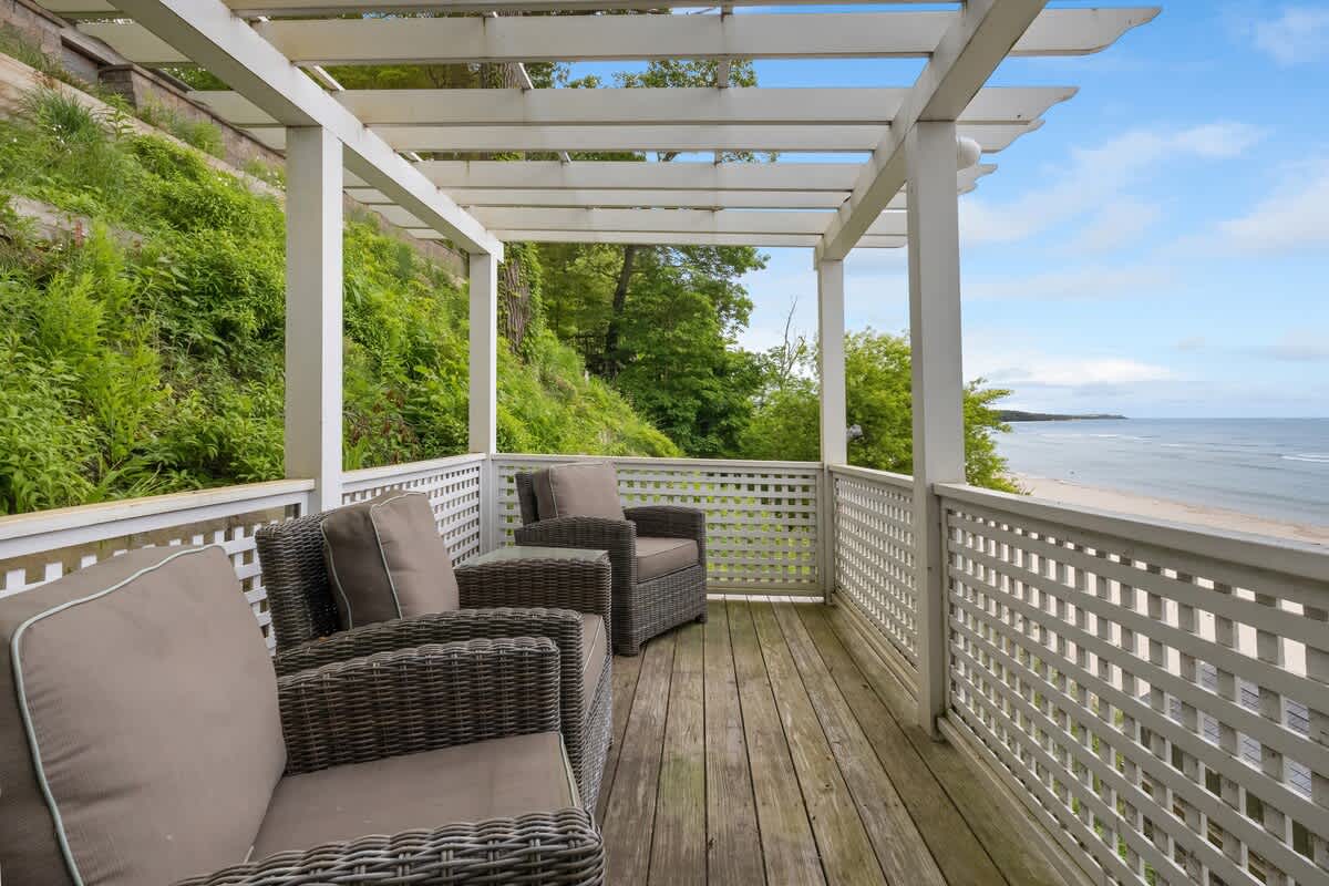 An elevated wooden deck, covered by a white pergola, features three comfortable outdoor armchairs. It offers a panoramic view of a sandy beach and the expansive blue ocean under a partly cloudy sky.