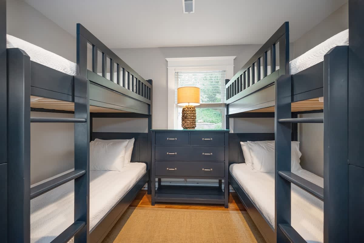 Kids' bedroom featuring two dark blue wooden bunk beds with crisp white bedding, flanking a matching dark blue dresser with a woven lamp. A window is centered above the dresser, and a light brown rug covers the floor.