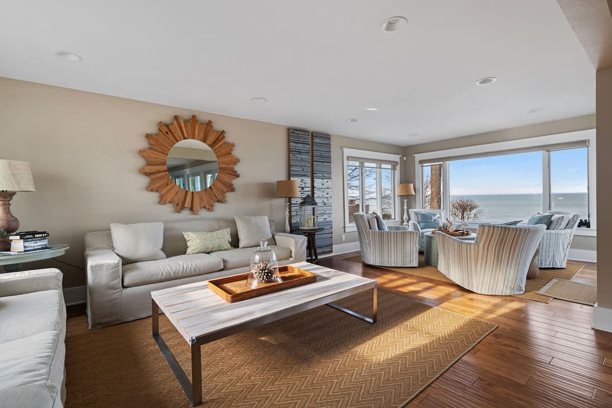 Spacious living room featuring a light-colored sofa, a wooden coffee table on a patterned rug, and a sunburst mirror, with a separate seating area overlooking the lake.