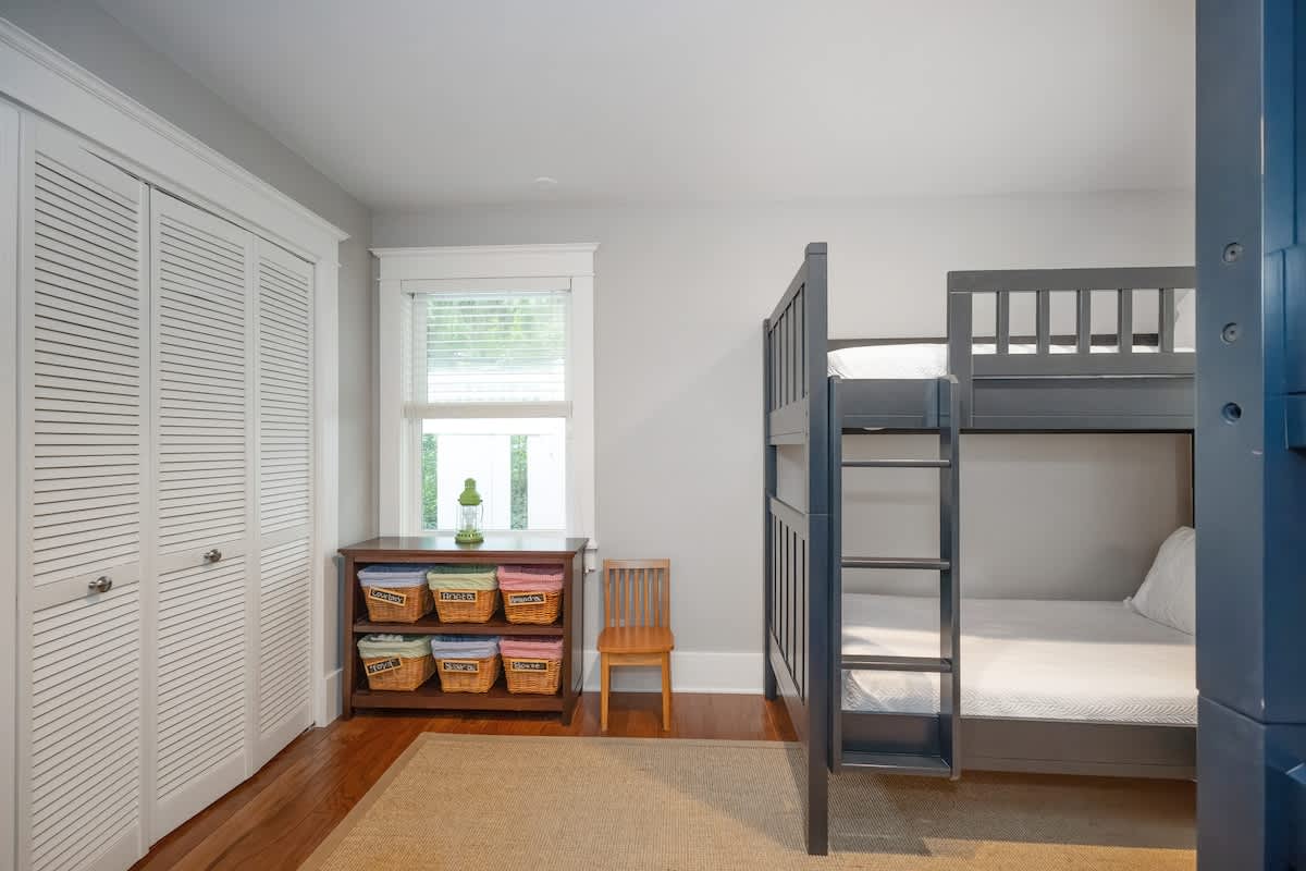 Kids' bedroom with a dark blue wooden bunk bed on the right and a white louvered closet on the left. A wooden shelf unit with woven baskets sits under a window, next to a small wooden chair, all on hardwood floors with a rug.