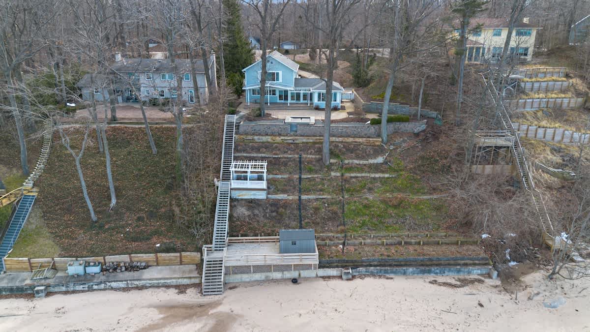An aerial view showcases the light blue lakefront house nestled among trees, with multiple long staircases descending the wooded slope to a sandy beach and the lake.