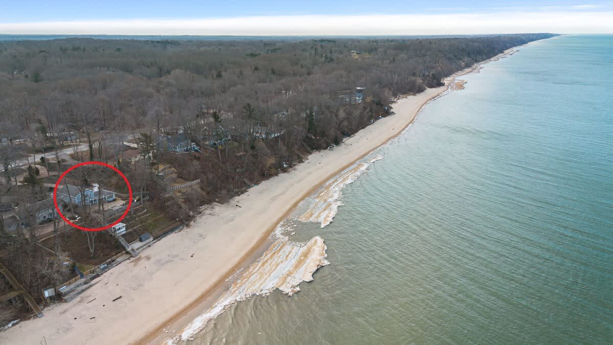 Wide aerial view of the coastline showing the property (highlighted by a red circle) situated on a bluff overlooking a sandy beach and a large body of water with ice formations.