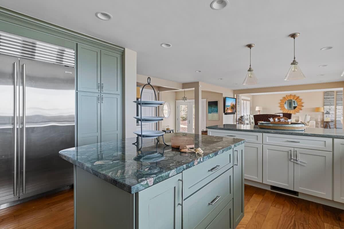 Kitchen area featuring a large stainless steel refrigerator and light green shaker cabinets with a matching green granite countertop on the island. A tiered serving stand sits on the island, connecting to other living spaces.