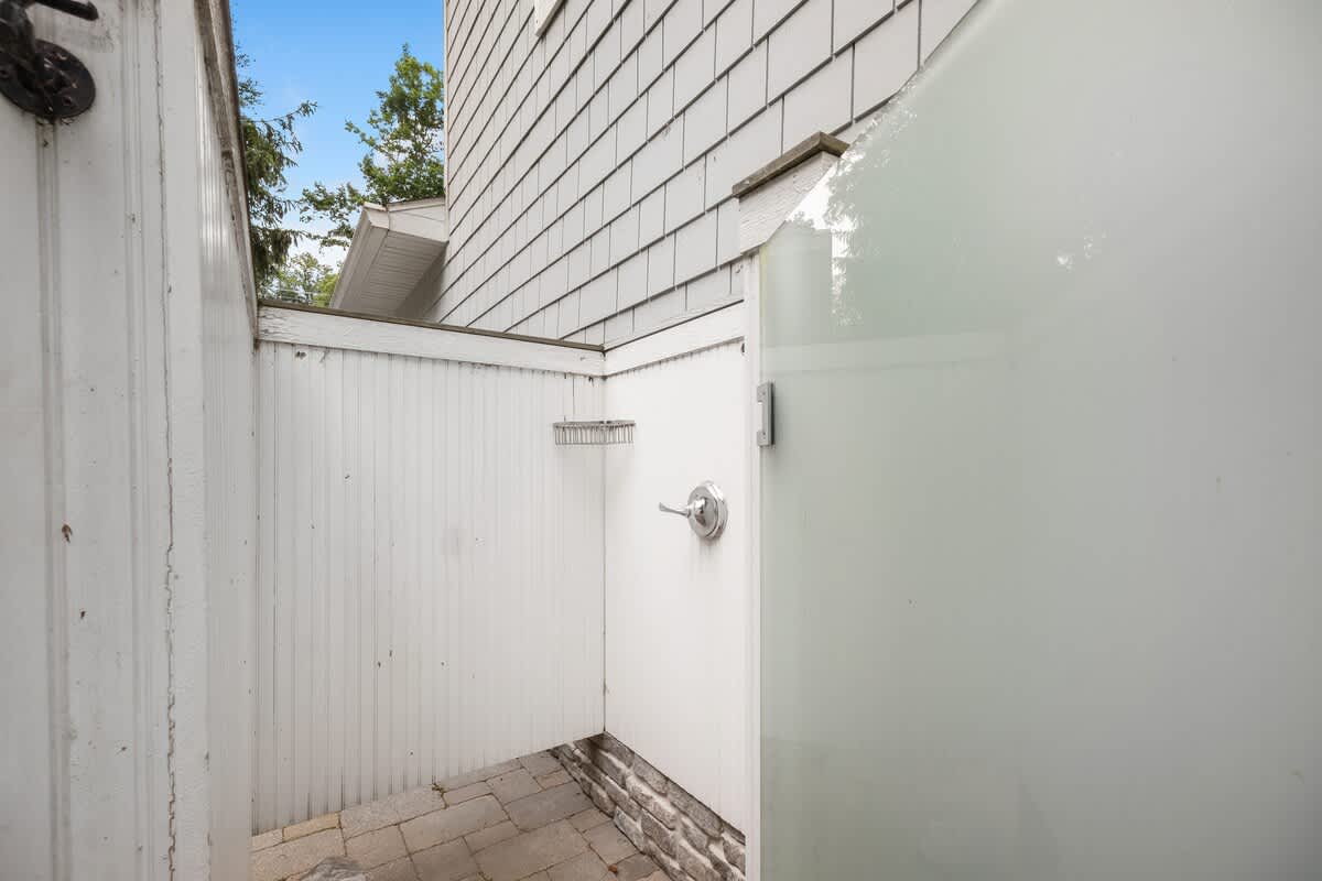 A private outdoor shower area features white paneled walls and a frosted glass door, with a simple shower fixture and a small shelf for toiletries. The floor is paved with light-colored bricks.