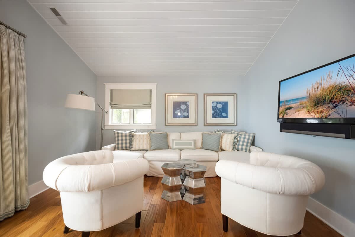 Bright living room with light blue walls, a white sectional sofa, and two white barrel chairs facing a pair of silver geometric coffee tables. A wall-mounted TV is on the right, and two framed blue botanical prints hang above the sofa, with a white shiplap ceiling.