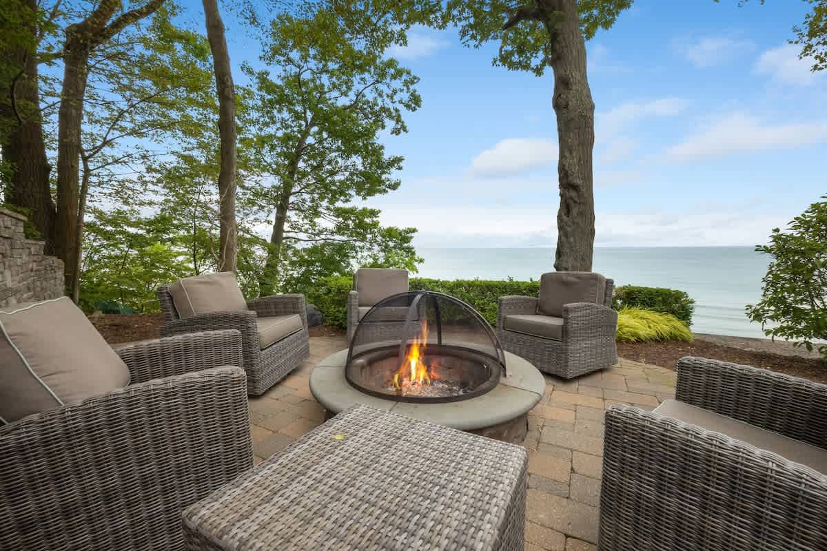 Cozy outdoor fire pit area with comfortable wicker armchairs arranged around a stone fire pit, set amidst trees with a serene lake backdrop.