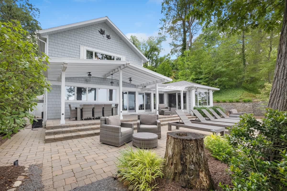 The back of a light gray shingle-sided house features a spacious paver patio with an outdoor dining set under a white pergola and several lounge chairs. Lush green trees and shrubs surround the property.