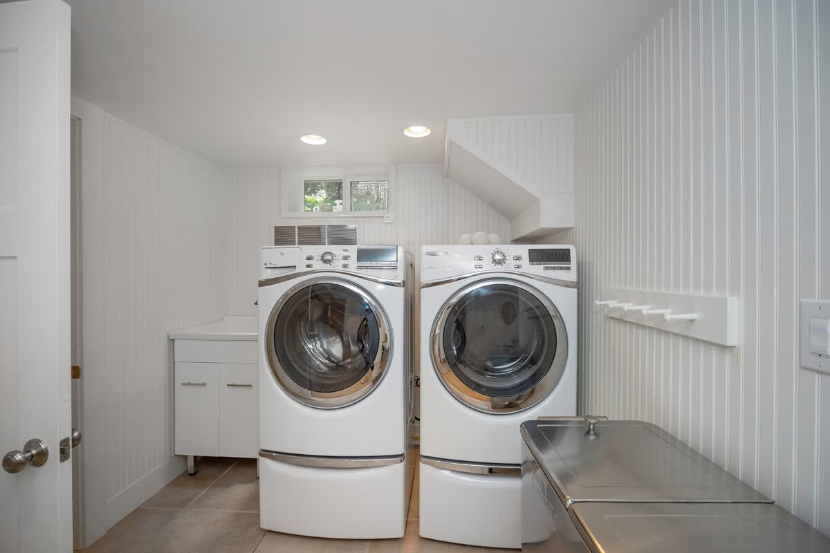 Bright and functional laundry room with side-by-side white front-loading washer and dryer on pedestals, a utility sink, and a wall-mounted drying rack.