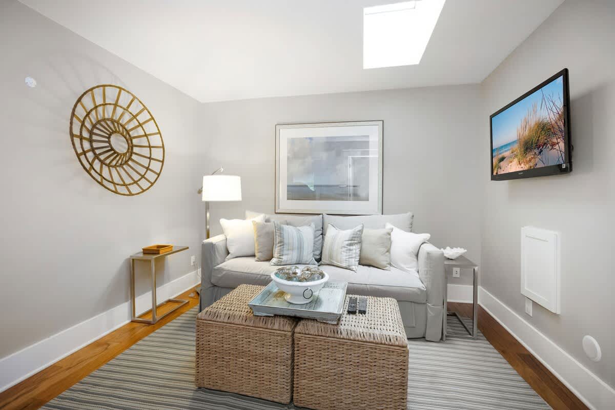 Cozy living area with light gray walls, a comfortable gray sofa with throw pillows, and a striped rug. Features include a wall-mounted TV, a skylight, and a decorative gold wall art piece.