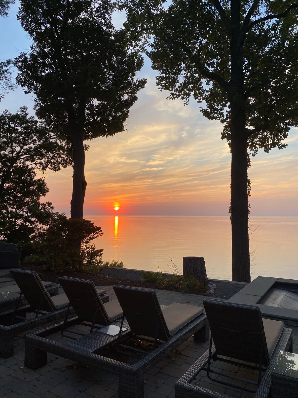 A serene sunset over a calm body of water, casting an orange glow, viewed from a paved patio with several dark lounge chairs. Two large trees frame the picturesque scene.