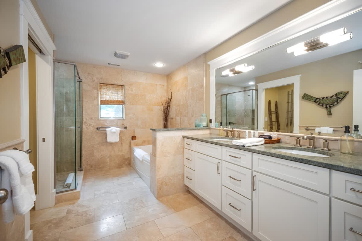 View of the beige tiled bathroom, showcasing a glass-enclosed walk-in shower next to the white soaking tub. The double vanity with its granite countertop and large mirror is also visible.