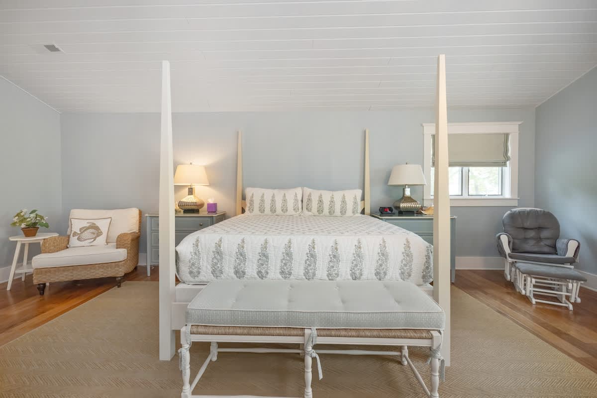 Serene bedroom with light blue walls, a white four-poster king-sized bed with patterned bedding, and hardwood floors. A light-colored rug anchors the bed, and a wicker chair sits in the corner.