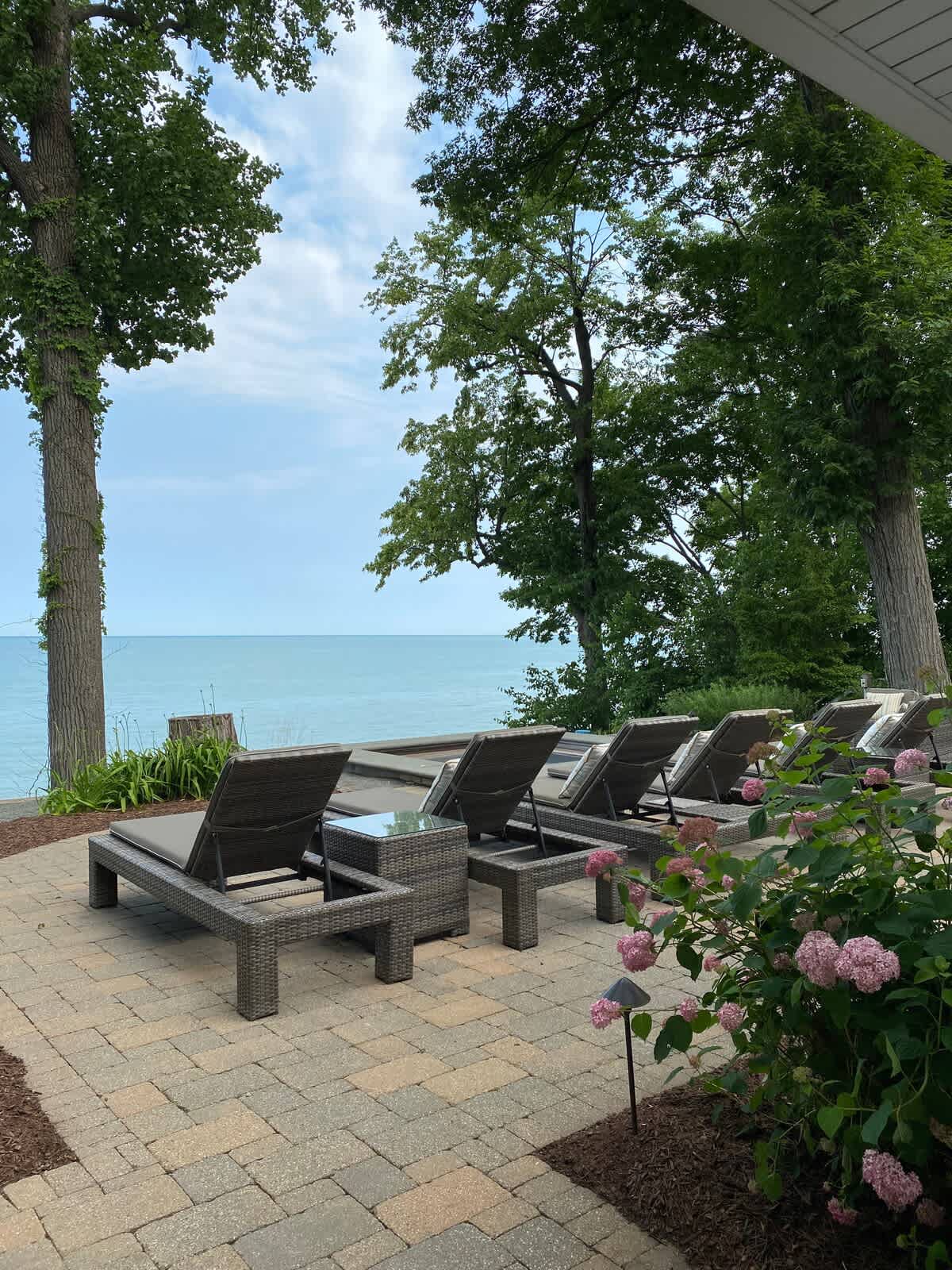 Expansive outdoor patio with several dark wicker lounge chairs arranged on pavers, offering direct, unobstructed views of the calm lake and lush trees.