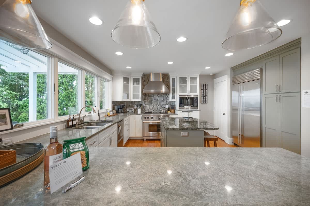 A modern kitchen features light gray granite countertops, white upper and light gray lower cabinetry, stainless steel appliances including a large refrigerator and range hood, and a central island with seating.