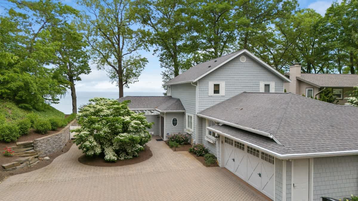 Front exterior of a light gray shingle-sided house with a paved driveway, an attached garage with white doors, and lush green landscaping with mature trees.