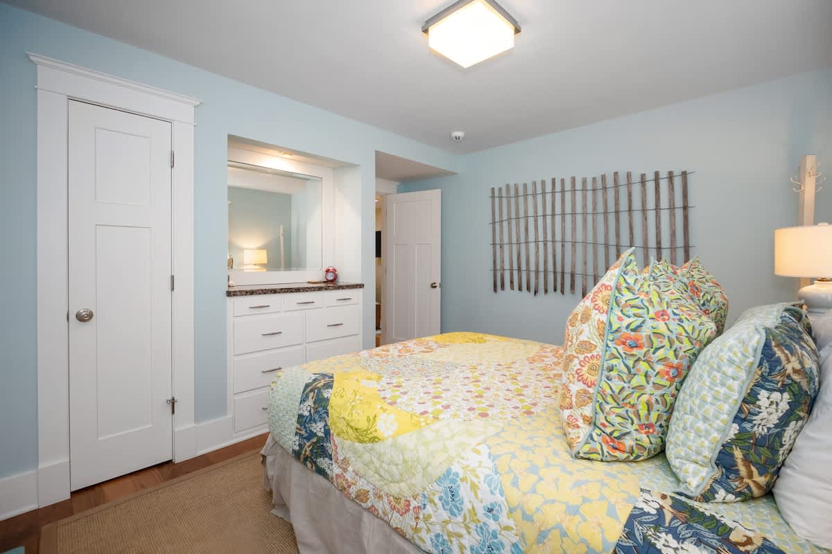 Colorful bedroom with a queen-sized bed, a patchwork quilt, and light blue walls. A built-in dresser with a mirror is visible on the left, and a decorative wooden wall art piece hangs on the right wall, adding a rustic touch.