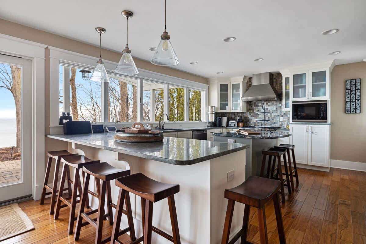 The bright kitchen boasts a long breakfast bar with wooden stools, light gray granite countertops, and a wall of large windows offering a glimpse of the lake, complemented by pendant lighting and hardwood floors.