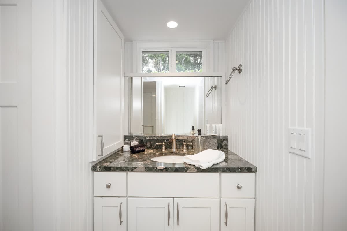 Bright white bathroom featuring beadboard walls, a dark granite countertop vanity with a single undermount sink, a large mirror, and a small window above.