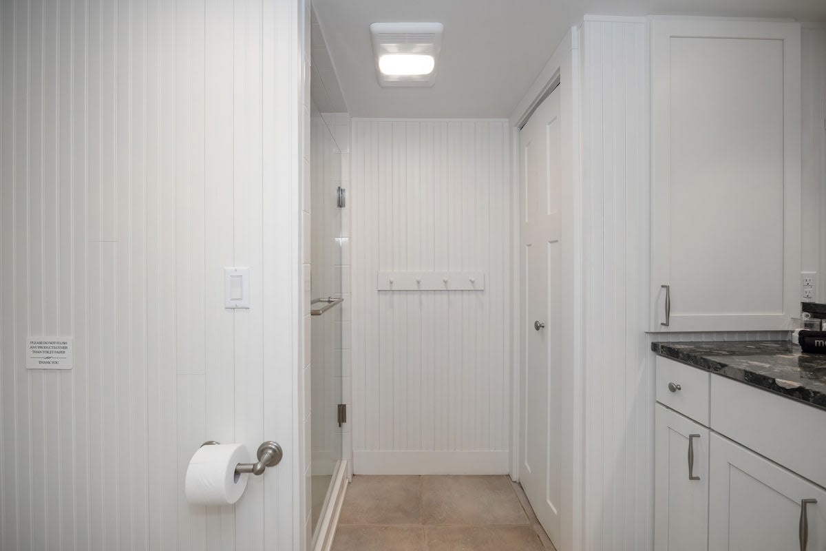 Clean white bathroom with beadboard walls, a glass-enclosed shower, a toilet with a wall-mounted toilet paper holder, and a small white coat rack.