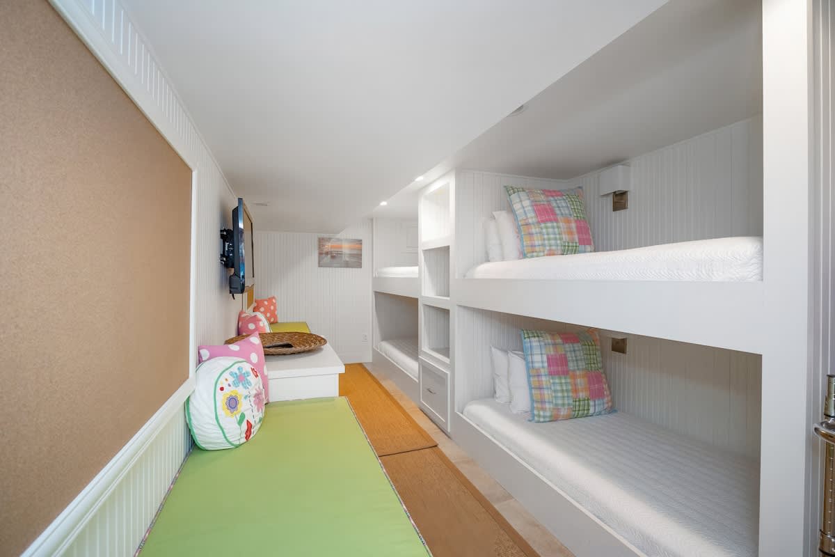 Long, narrow bunk room with built-in white bunk beds featuring colorful plaid pillows, a long green cushioned bench with storage, and a wall-mounted TV.