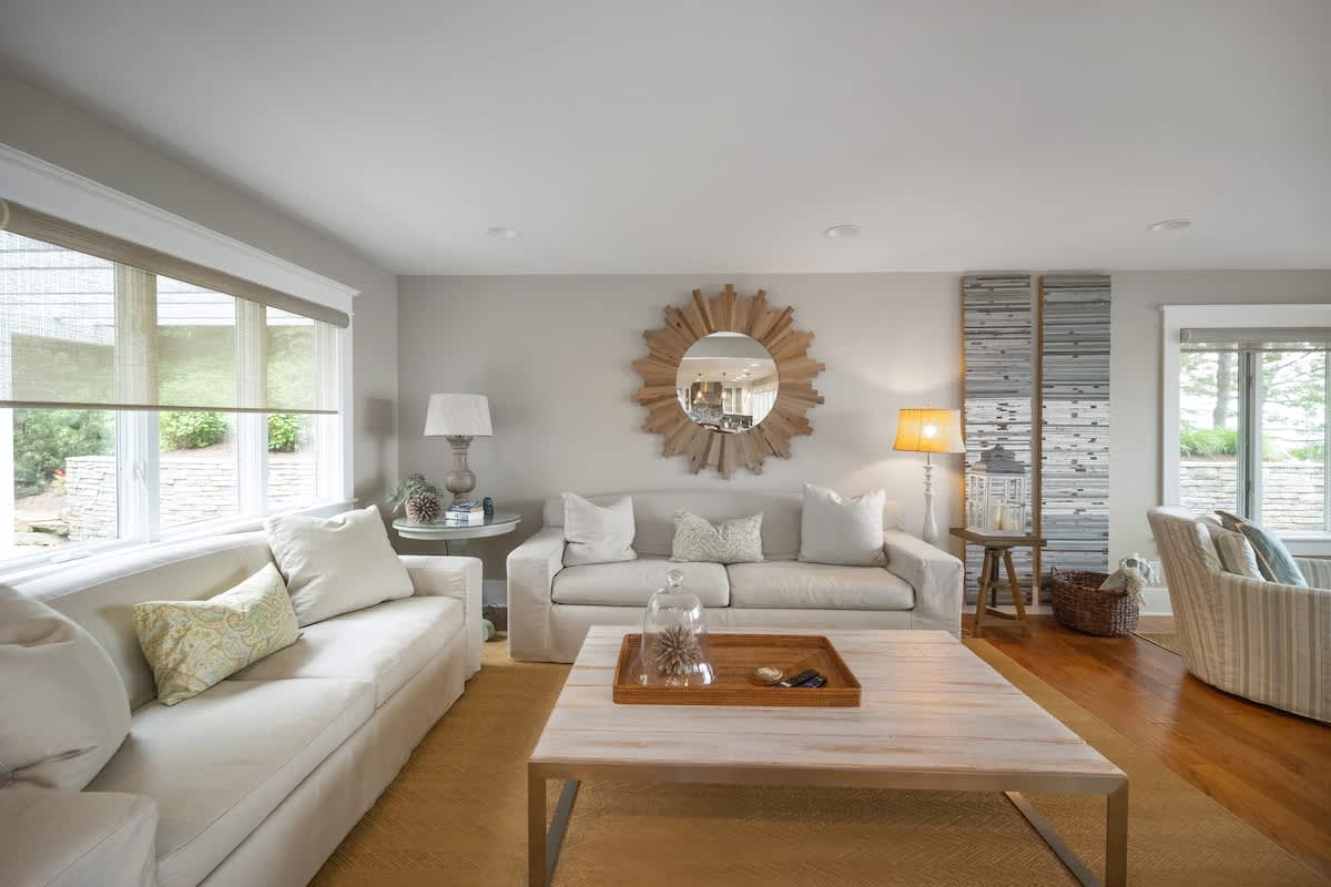 The main living room boasts light-colored walls, hardwood floors, a large white slipcovered sofa, a light wood coffee table with a decorative tray, and a prominent sunburst mirror above another sofa.