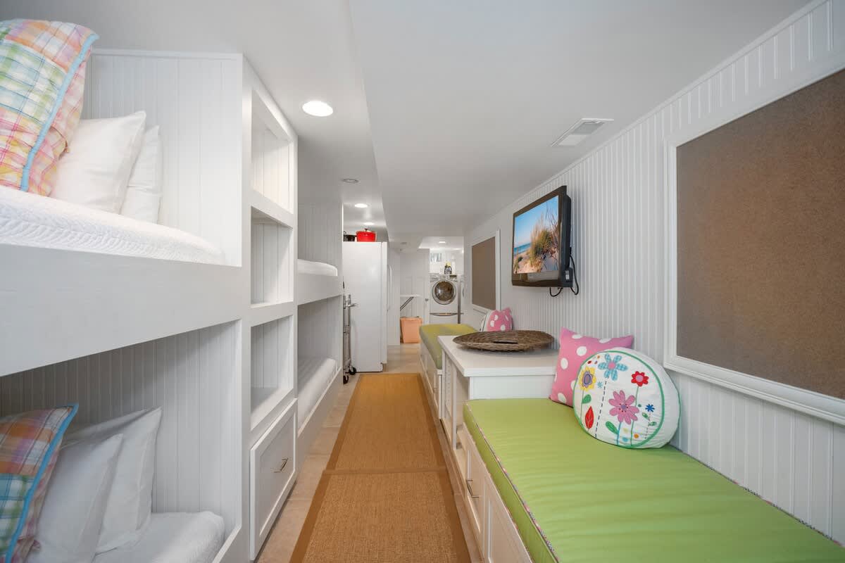 Another view of the bunk room, showcasing built-in white bunk beds, a green cushioned bench, and a wall-mounted TV, with a laundry area visible in the background.
