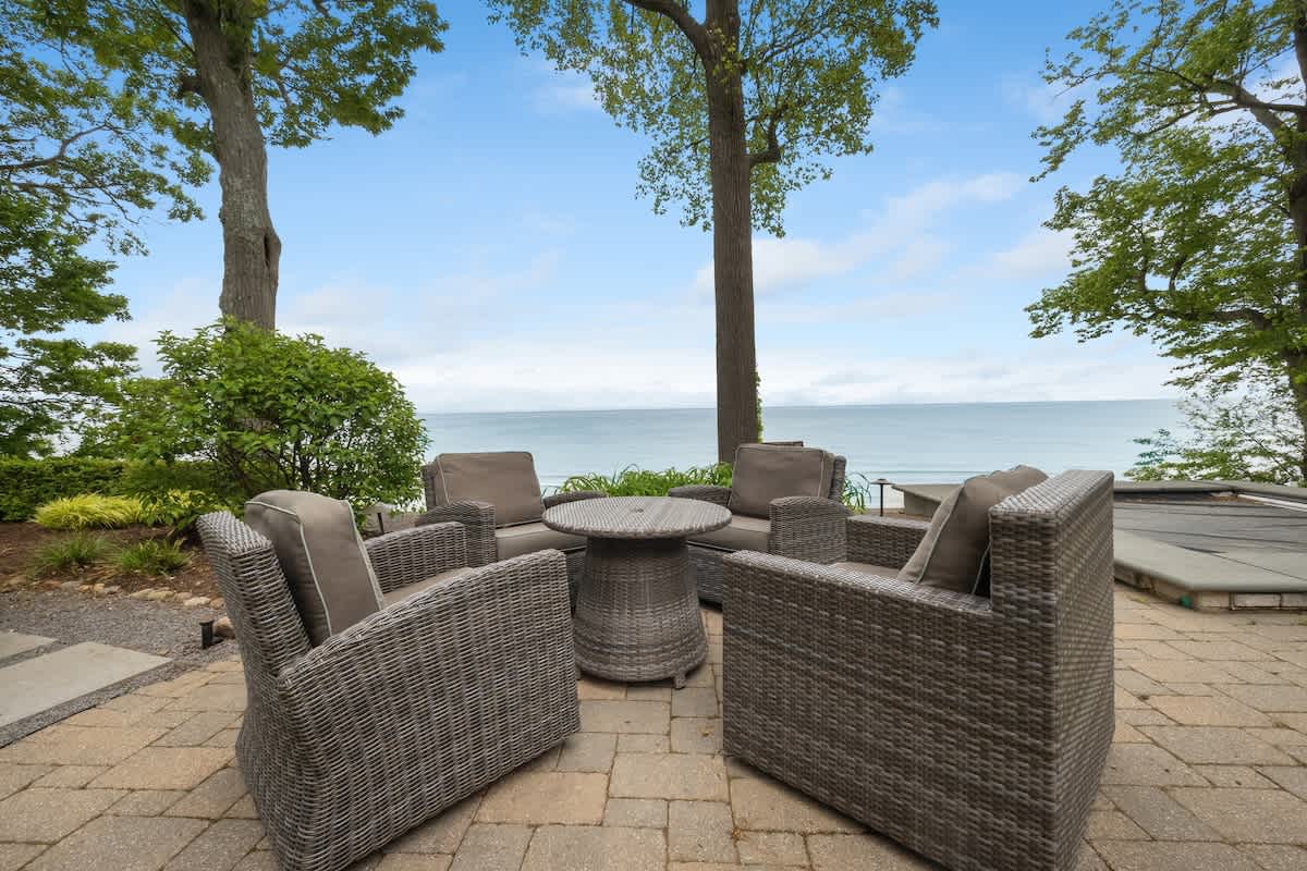Outdoor paver patio with gray wicker chairs and a round table, offering a serene view of a large body of water and lush green trees under a clear sky.