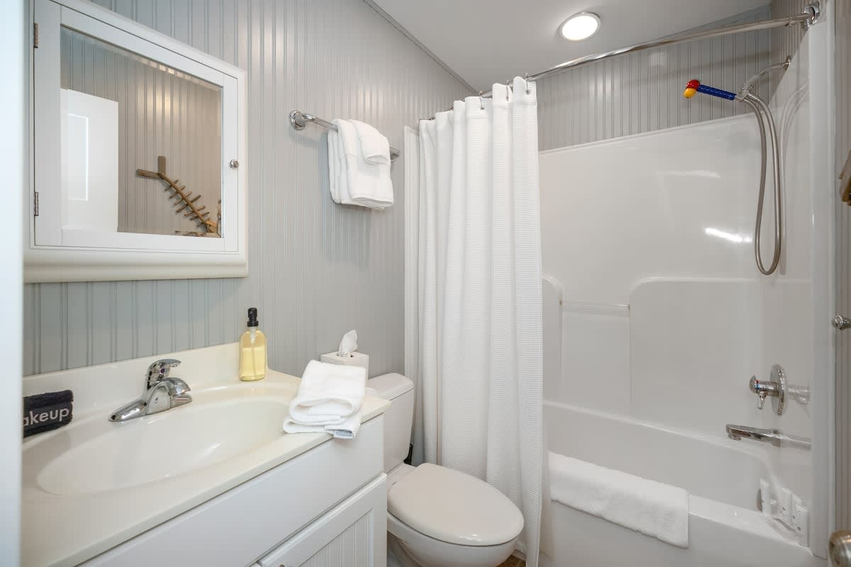 Compact bathroom with white beadboard walls, a white vanity with a sink, toilet, and a white shower/tub combo with a white curtain. A mirrored medicine cabinet hangs above the sink, providing storage.